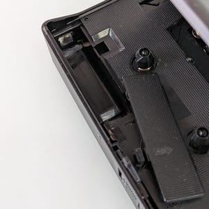 May include: A black vintage cassette player with the top open, revealing the internal components. The interior features a textured surface and various mechanical parts. The player's design is simple and functional, with a focus on the mechanics of the device.