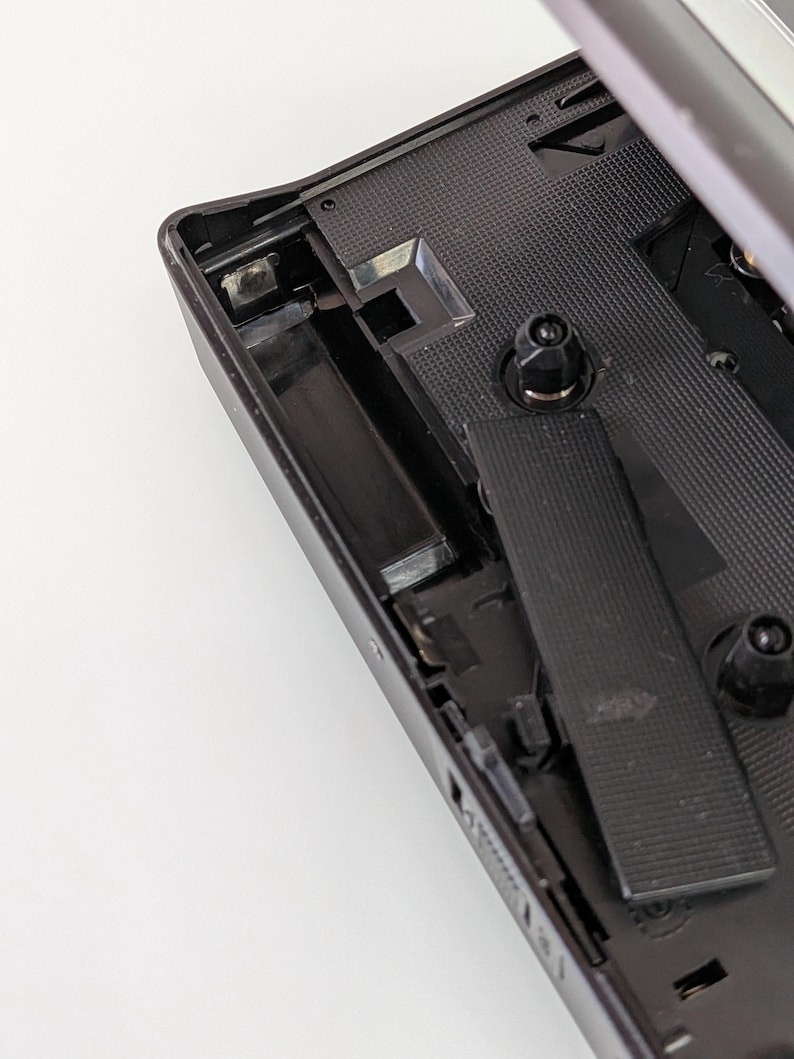 May include: A black vintage cassette player with the top open, revealing the internal components. The interior features a textured surface and various mechanical parts. The player's design is simple and functional, with a focus on the mechanics of the device.