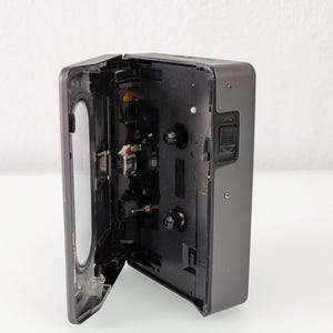 May include: A dark grey cassette player with the cassette door open, showing the internal mechanisms. The player has a rectangular shape with rounded edges and a silver hinge. The "OPEN" button is visible on the side of the device.