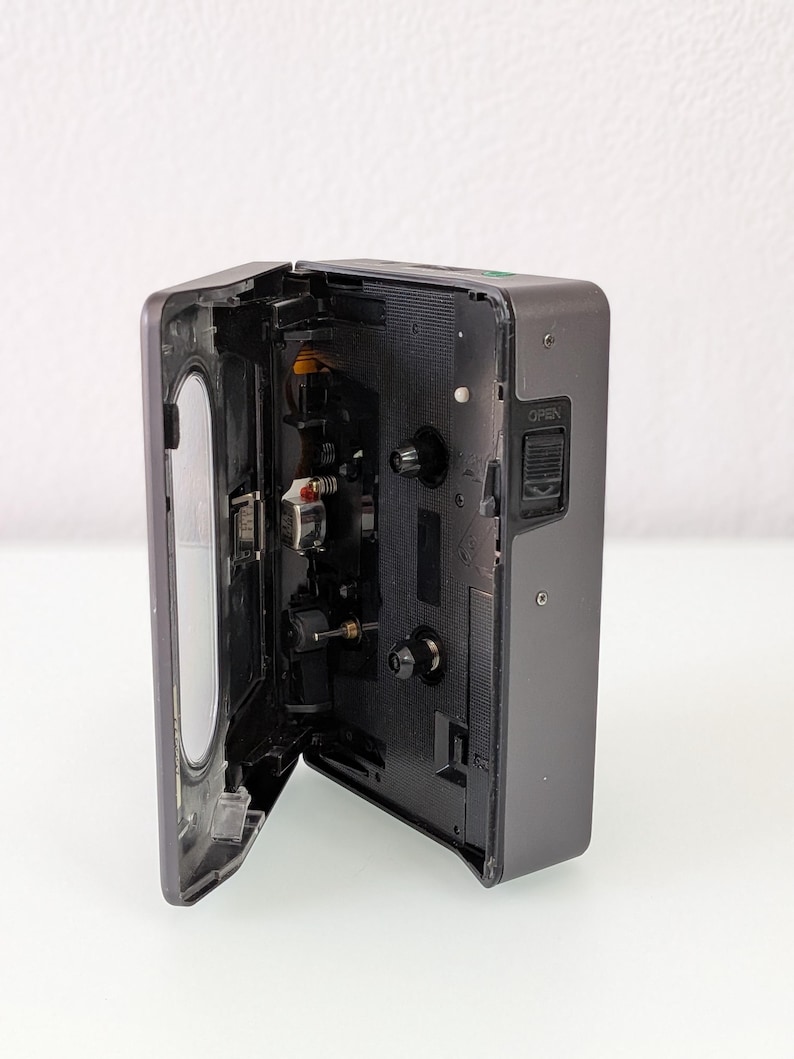 May include: A dark grey cassette player with the cassette door open, showing the internal mechanisms. The player has a rectangular shape with rounded edges and a silver hinge. The "OPEN" button is visible on the side of the device.