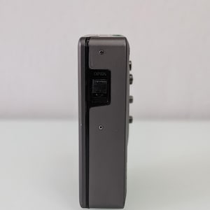 May include: A grey, rectangular audio cassette player with a black control panel. The word "OPEN" is visible on the side. The device has several small buttons and a small circular hole. The device is on a white background.