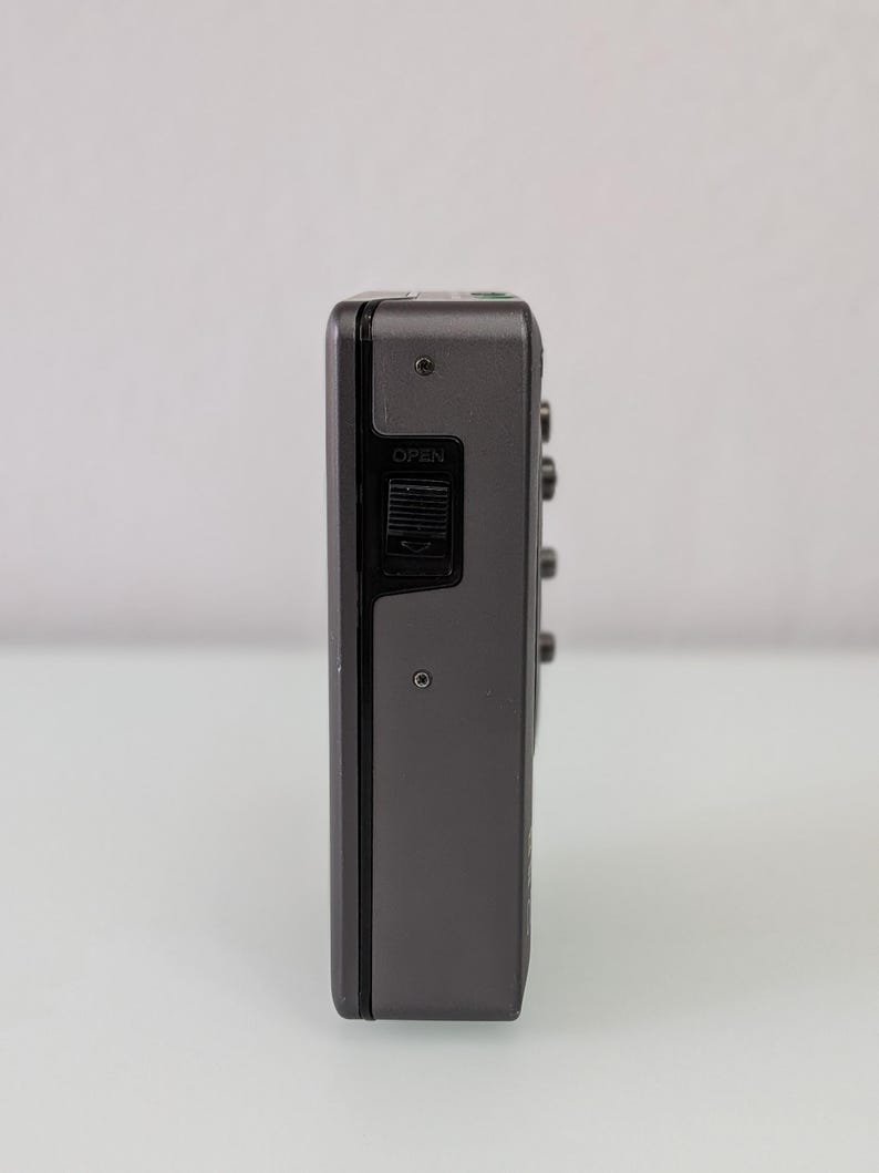 May include: A grey, rectangular audio cassette player with a black control panel. The word "OPEN" is visible on the side. The device has several small buttons and a small circular hole. The device is on a white background.