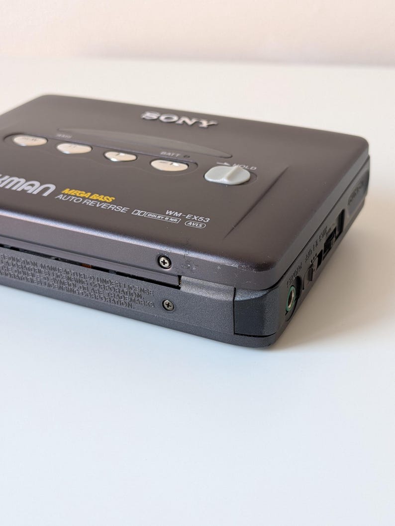 May include: A dark gray Sony Walkman WM-EX53 cassette player featuring Mega Bass, Auto Reverse, and Dolby B NR technology. The device includes silver control buttons, a hold switch, and a headphone jack on the side, with printed model details.