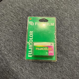 Puede incluir: Un paquete de película Fujifilm Fujicolor 200 para impresiones en color. La película está en un paquete de plástico verde y transparente. El cartucho de película es verde con las palabras "Fujicolor 200" en blanco y rosa. El paquete también tiene el texto "36".