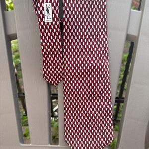 Könnte beinhalten: Eine weinrote Krawatte mit einem sich wiederholenden weißen Rautenmuster. Die Krawatte hat eine schwarze Rückseite und ein Etikett mit dem Text "YSL PARIS". Die Krawatte ist über einen grauen Stuhl drapiert.