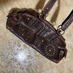 May include: A dark brown leather handbag with decorative stitching and brass-colored studs. The bag features a shoulder strap and a circular design on the front. The bag is a satchel style.
