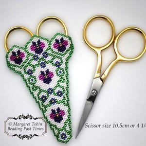 Könnte beinhalten: Perlenbesetzter Scherenhalter mit einem grünen, weißen und violetten Blumendesign. Der Halter ist aus kleinen Perlen gefertigt und zeigt ein zartes Blumenmuster. Der Halter ist für eine Schere konzipiert, die auf dem Bild zu sehen ist. Die Schere ist 10,5 cm lang.