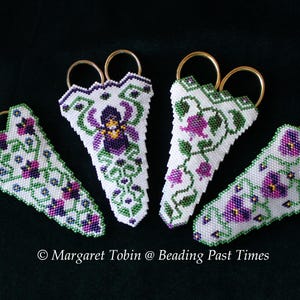 May include: Four white beaded scissor fobs with gold-colored rings, each featuring a different floral design in purple, green, and white. The fobs are arranged against a dark green background. Text: Margaret Tobin @ Beading Past Times.