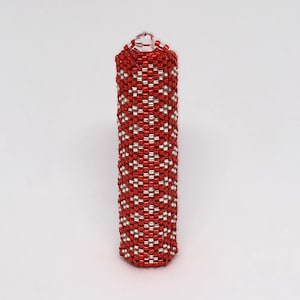 Puede incluir: Un objeto cilíndrico de cuentas, principalmente rojo con un patrón repetitivo de cuentas plateadas. La parte superior tiene un pequeño lazo. El objeto parece ser un recipiente o una pieza decorativa. Las cuentas son pequeñas y están tejidas apretadamente.
