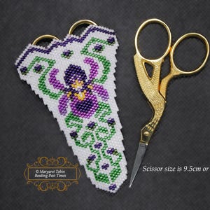 Może przedstawiać: Białe koralikowe etui na nożyczki z fioletowym i zielonym wzorem kwiatowym. Etui jest przeznaczone do przechowywania małych złotych nożyczek o długości 9,5 cm. Tekst "Margaret Tobin Beading Past Times" jest widoczny w lewym dolnym rogu obrazu.