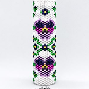 May include: A cylindrical beaded tube featuring a white background with a repeating pattern of purple and dark blue hearts, green accents, and small yellow centres. The top is finished with a faceted dark purple crystal bead.