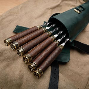 Può includere: Un set di sei spiedini in metallo con manici in legno, tutti in una custodia di trasporto in pelle verde. Gli spiedini hanno una punta in ottone decorativa e sono disposti nella custodia con i manici che sporgono.