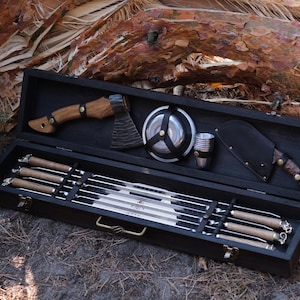 May include: A black wooden case containing a barbecue tool set. The set includes skewers, a small axe, a knife with a leather sheath, a metal bowl, and a small cup. The tools have wooden handles and are displayed on a bed of black felt.