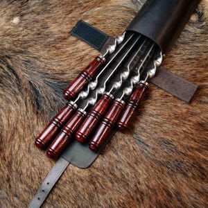 Pode incluir: Conjunto de seis espetos de aço inoxidável com cabos de madeira vermelha escura, guardados numa bainha de couro preta. Os espetos têm um design torcido e são ideais para grelhar. A bainha tem uma correia com fivela.