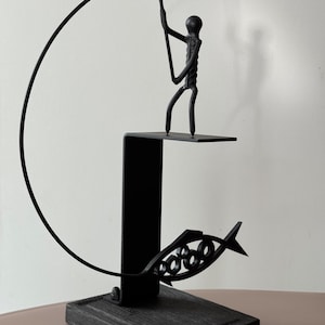 May include: Black metal sculpture of a fisherman with a fishing rod, standing on a platform. A stylized fish is below the platform. The sculpture is mounted on a dark, rectangular wooden base. The fishing rod curves in an arc.