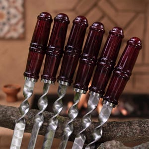 May include: A set of six metal skewers featuring dark reddish-brown wooden handles with a cylindrical, ribbed design. The metal shafts have a distinct twisted, spiral pattern leading to a flat, polished blade, providing a sturdy grip for cooking.
