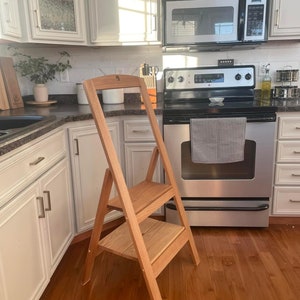 May include: A wooden step stool with two steps. The stool is made of light brown wood and has a natural finish. It is standing in a kitchen with white cabinets and a stainless steel stove.