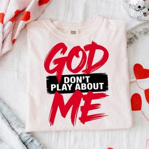 May include: Cream-colored t-shirt with the text "GOD DON'T PLAY ABOUT ME" in bold red lettering. The words "DON'T PLAY ABOUT" are in white text on a black horizontal rectangle. The shirt is folded on a white surface.
