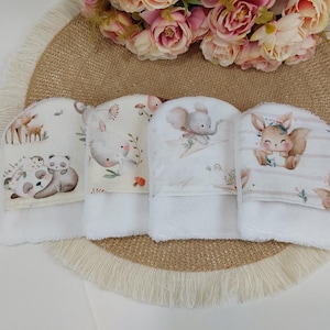 Puede incluir: Cuatro toallitas blancas con ilustraciones de animales, incluyendo un panda, un ciervo, un elefante y una ardilla. Cada toallita tiene un diseño de animal impreso en la capucha y una base de rizo blanca. Las toallitas están dispuestas sobre una estera tejida, con flores en el fondo.