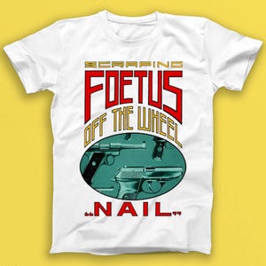 Puede incluir: Camiseta blanca con un diseño gráfico. El diseño presenta el texto "SCRAPING FOETUS OFF THE WHEEL" en letras rojas y doradas, junto con dos ilustraciones de pistolas dentro de un óvalo turquesa y la palabra "NAIL" en rojo.