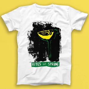 Peut inclure: T-shirt blanc avec un motif graphique d'une fleur jaune sur fond noir. Le design comprend le texte "RITES OF SPRING" dans un rectangle vert. Le t-shirt est en matière souple, idéal pour un usage quotidien.