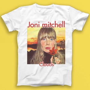 May include: White t-shirt featuring a portrait of Joni Mitchell with the text "Joni Mitchell" in red above the image and "Clouds" in red below. The portrait shows a woman holding a red flower against a sunset backdrop.