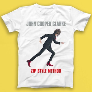 May include: White t-shirt featuring a black and white image of a man in a suit and red shoes. The text "John Cooper Clarke" and "Zip Style Method" is printed on the shirt.
