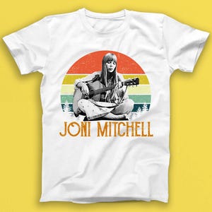 May include: White t-shirt with a vintage-style illustration of Joni Mitchell playing a guitar in front of a sunset. The text "Joni Mitchell" is printed below the image.