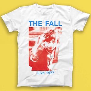 May include: White t-shirt featuring a red and white image of a man making a peace sign. The text "THE FALL" is in blue above the image and "Live 1977" is in blue below the image.