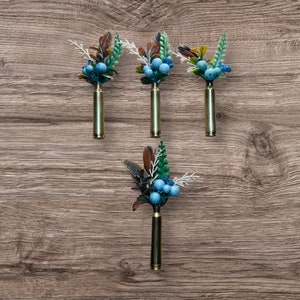 May include: Four decorative boutonnieres. Each boutonniere features a brass bullet casing base, topped with a cluster of blue berries, green and white sprigs, and brown feathers. The items are arranged on a wooden surface.