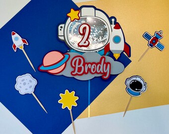 May include: A set of cake toppers featuring an astronaut helmet, a rocket, a planet, a star, a satellite, and a cloud with the name "Brody" on it. The astronaut helmet has a number "2" on it.