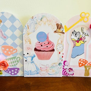 May include: Three Alice in Wonderland themed party decorations. The first is blue and white with a checkered pattern and a bottle with the text "Drink Me". The second is white with a cupcake and the text "Eat Me". The third is pink with a keyhole and a cartoon image of the Mad Hatter.