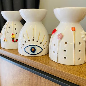May include: Three white ceramic oil warmers with decorative designs. One features a gold and blue eye, another has Christmas-themed charms, and the third has floral accents. Each warmer has a wide top and a series of holes.