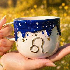 May include: A white ceramic mug with a gold handle and the Leo zodiac symbol. The top of the mug is decorated with a blue, starry design. The word "Leo" is written in blue with gold accents.