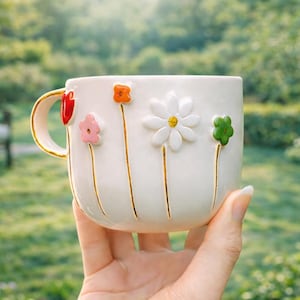May include: White ceramic mug with a gold handle and floral embellishments. The mug features colourful, raised flower designs in red, orange, pink, white, and green. The flowers are attached to the mug with gold stems.