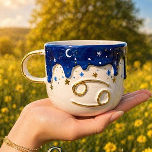 May include: A white ceramic mug with a gold handle and the Cancer zodiac symbol. The mug features a blue rim with a starry night design and the word "Cancer" in gold lettering.