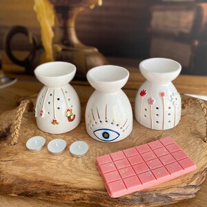 May include: Three white ceramic oil burners with distinct designs: one with hanging Christmas ornaments, one with a central blue eye motif, and one with small floral accents. Includes three small white tea light candles and a pink wax melt bar.