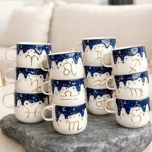 May include: A collection of ceramic mugs featuring a white base and a dark blue drip pattern at the top, decorated with gold stars and raised gold zodiac symbols. Each mug has a side handle and displays a unique astrological sign in gold.