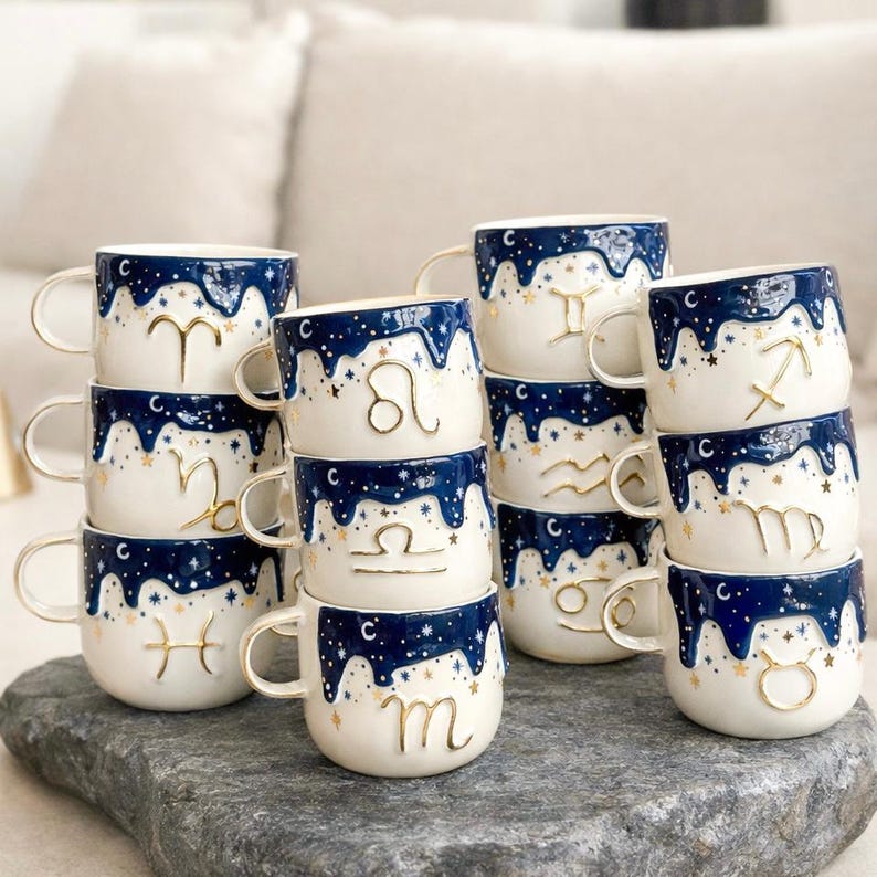 May include: A collection of ceramic mugs featuring a white base and a dark blue drip pattern at the top, decorated with gold stars and raised gold zodiac symbols. Each mug has a side handle and displays a unique astrological sign in gold.