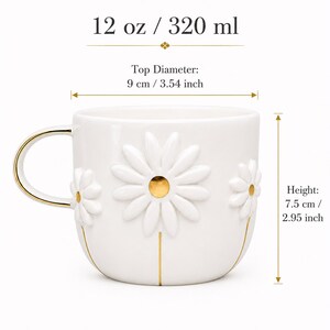 May include: A white ceramic mug with embossed daisy flowers featuring gold centers and stems. The mug has a gold-trimmed handle, a 12 oz capacity, a 3.54 inch top diameter, and a 2.95 inch height. Text indicates 12 oz / 320 ml, 9 cm / 3.54 inch, and 7.5 cm / 2.95 inch.