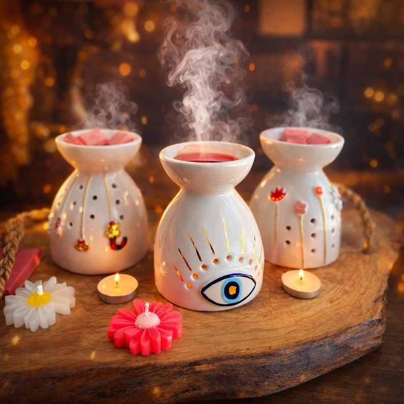 May include: Three white ceramic essential oil diffusers with decorative cutouts and gold accents, each holding a pink wax melt. A lit candle sits in front of each diffuser. Decorative candles shaped like flowers are also present.