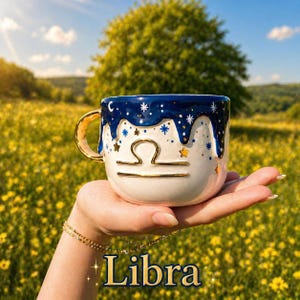 May include: A ceramic mug featuring a white base with a dark blue drip pattern at the top. The mug is decorated with gold stars and a gold Libra zodiac symbol on the front. It has a gold-colored handle and the word Libra printed at the bottom.