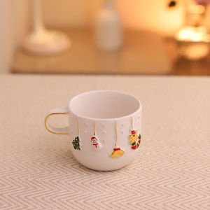 May include: White ceramic mug with a gold handle and gold trim. The mug is decorated with colorful Christmas ornaments hanging from gold strings. Ornaments include a Christmas tree, Santa, a bell, and a reindeer.