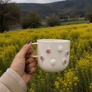 May include: A white ceramic mug with a gold handle and floral embellishments. The mug features raised, stylized flowers in white and pink, with gold accents. The mug is held in front of a field of yellow flowers.