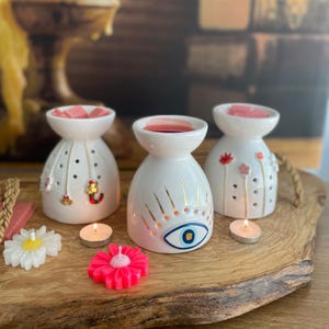 May include: Three white ceramic wax warmers with a bowl top and perforated sides. One features a blue and gold evil eye design, one has small hanging charms, and one displays painted floral stems. Includes two small tea light candles and two flower-shaped wax melts.