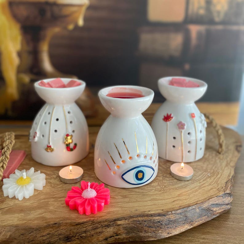 May include: Three white ceramic wax warmers with a bowl top and perforated sides. One features a blue and gold evil eye design, one has small hanging charms, and one displays painted floral stems. Includes two small tea light candles and two flower-shaped wax melts.