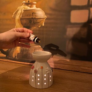 May include: A white ceramic oil burner with a flared top bowl and a base featuring small circular cutouts and colorful floral accents. The burner is positioned below a small amber glass dropper bottle containing essential oil.