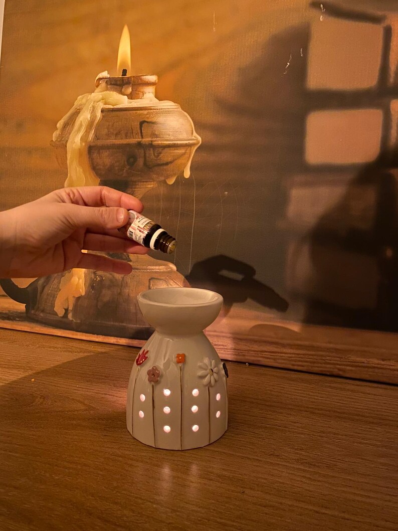 May include: A white ceramic oil burner with a flared top bowl and a base featuring small circular cutouts and colorful floral accents. The burner is positioned below a small amber glass dropper bottle containing essential oil.