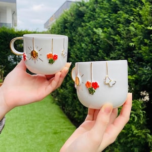 May include: Two white ceramic mugs with gold handles and decorative accents. One mug features a sun design, while both have floral and butterfly embellishments. The mugs are held against a backdrop of green foliage.
