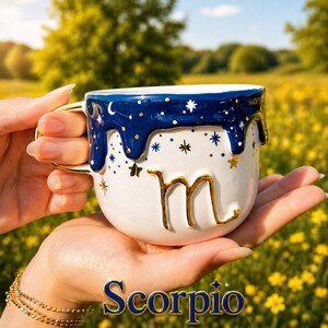 May include: A white ceramic mug featuring a dark blue drip glaze at the rim, decorated with gold and blue star motifs. The mug displays a prominent gold-colored letter m on the front and includes the word Scorpio in blue text at the bottom.